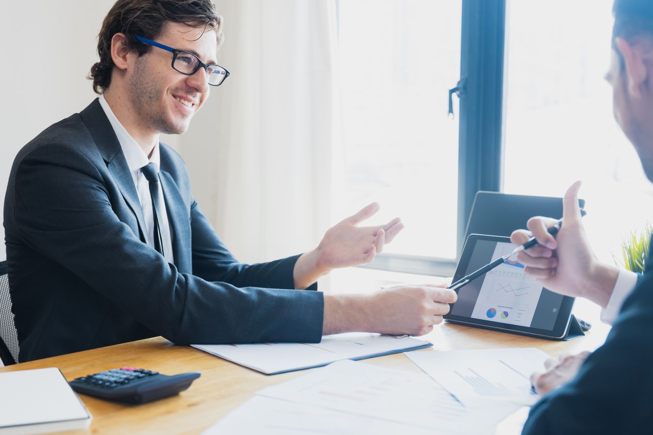 Two businessmen in formal attire discussing a business report in an office setting with a tablet.