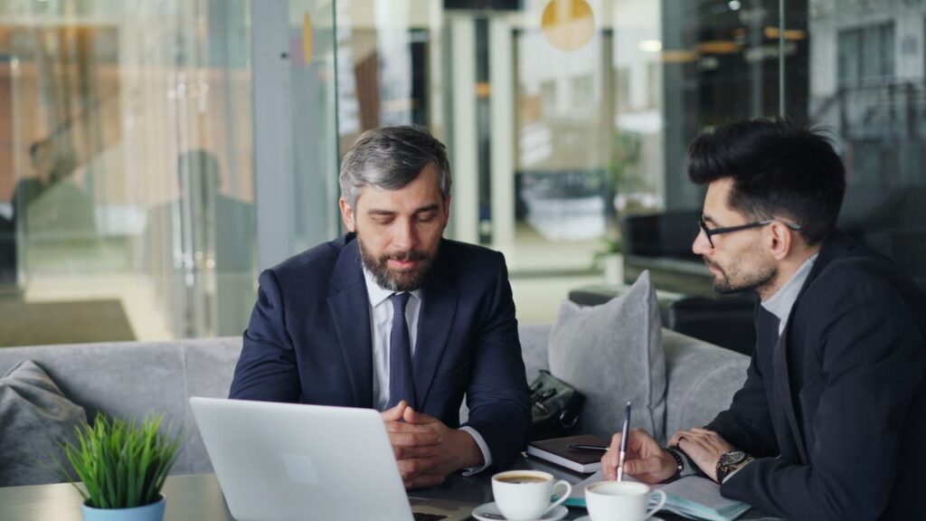 Two businessmen in suits discussing work at a cafe with laptops and coffee.