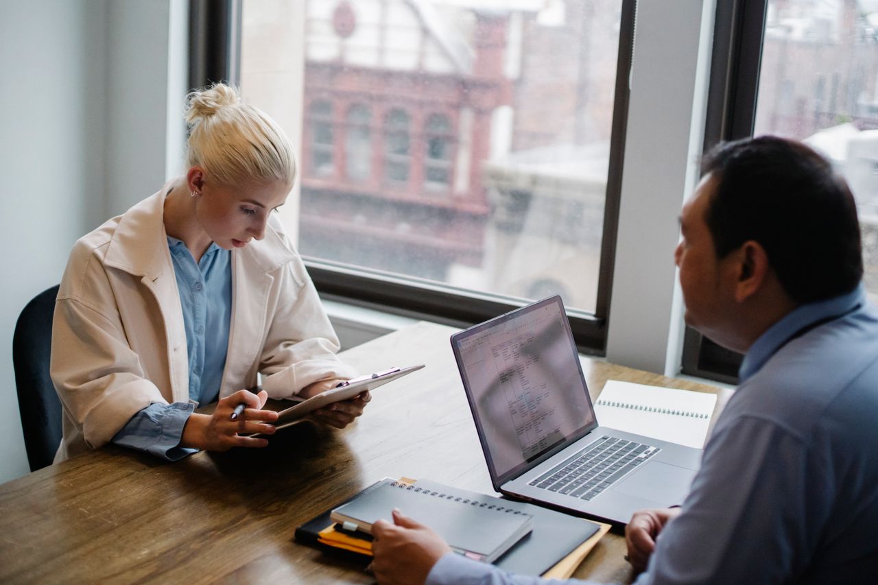 From above of young female worker wearing modern coat analyzing details of project with colleague while sitting together at wooden desk with laptop and documents
